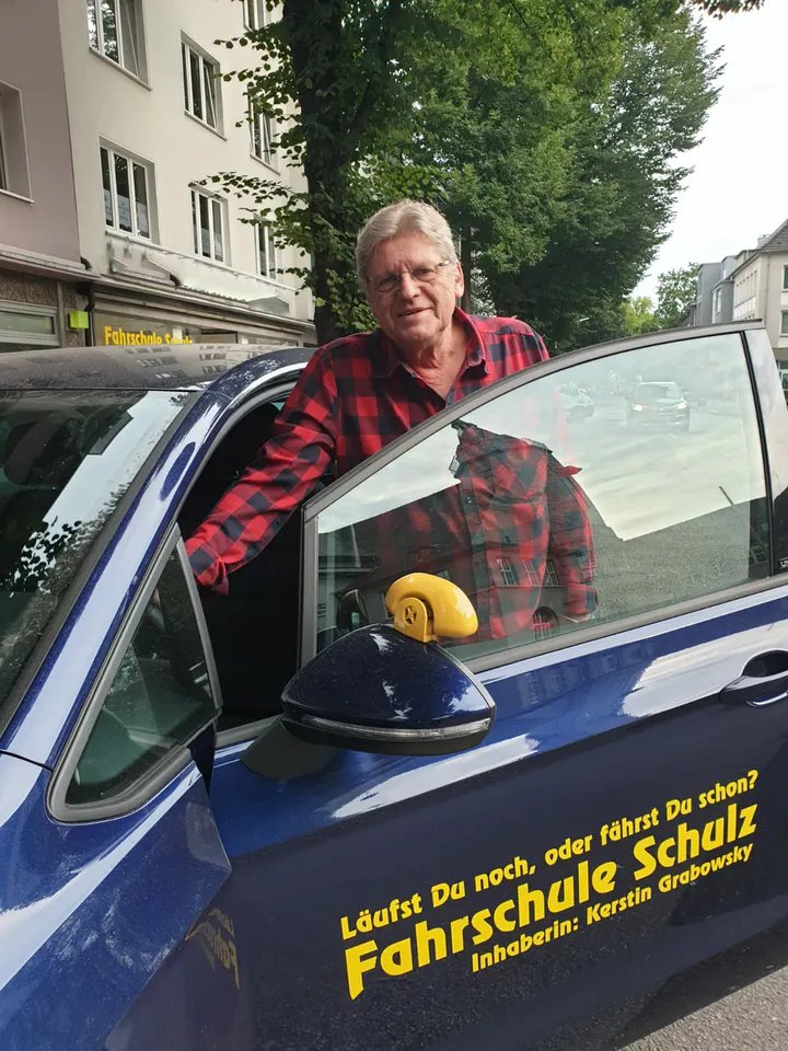 Fahrlehrer der Fahrschule Schulz an der geöffneten Tür des dunkelblauen Schulwagens, im Hintergrund die Ladenfront der Hauptstelle